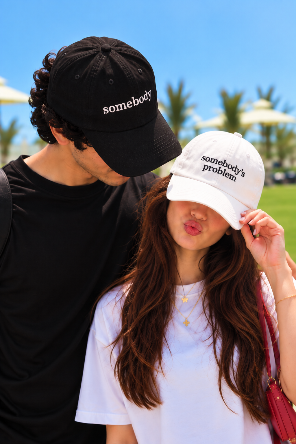 Two people outdoors wearing embroidered baseball caps. The man wears a black cap with 'somebody' in white text, and the woman wears a white cap with 'somebody's problem' in black text.